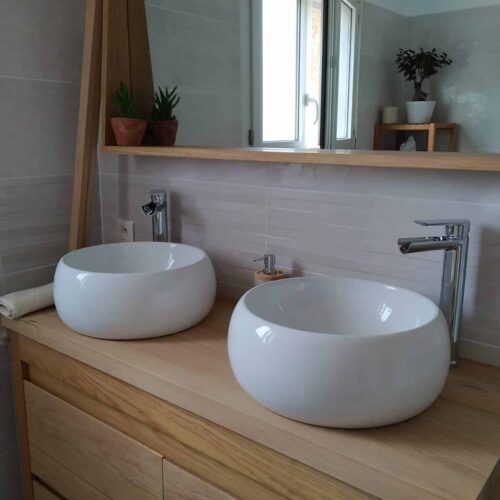 Salle de bain moderne avec deux vasques rondes blanches sur un meuble en bois, robinets chromés et fenêtre, étagère avec plantes et diffuseur.