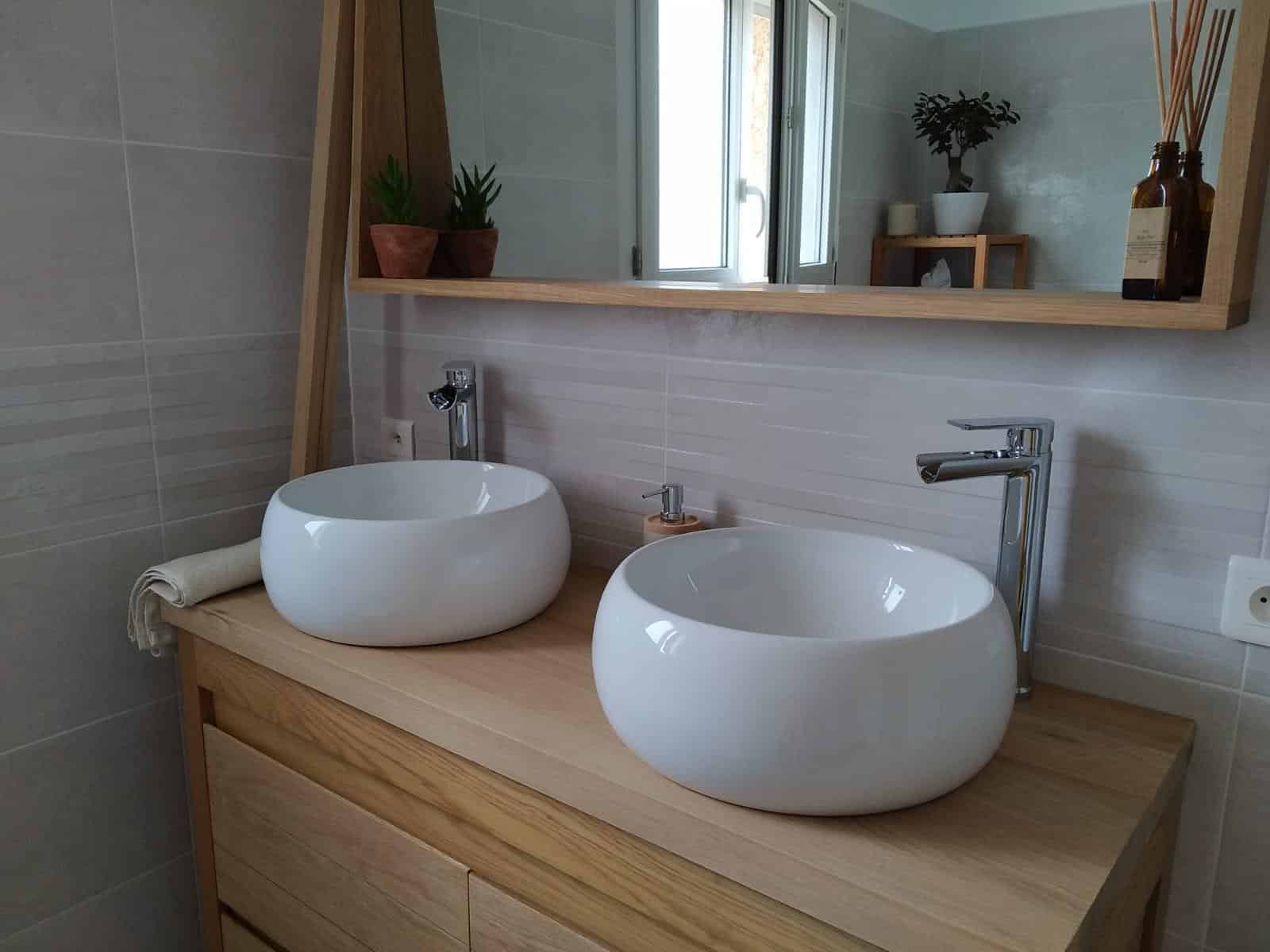 Salle de bain moderne avec deux vasques rondes blanches sur un meuble en bois, robinets chromés et fenêtre, étagère avec plantes et diffuseur.