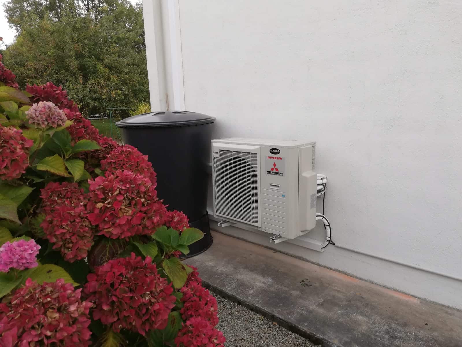 Un climatiseur extérieur Mitsubishi posé contre un mur blanc, près de grandes fleurs roses et rouges en façade et d’une citerne noire cylindrique.