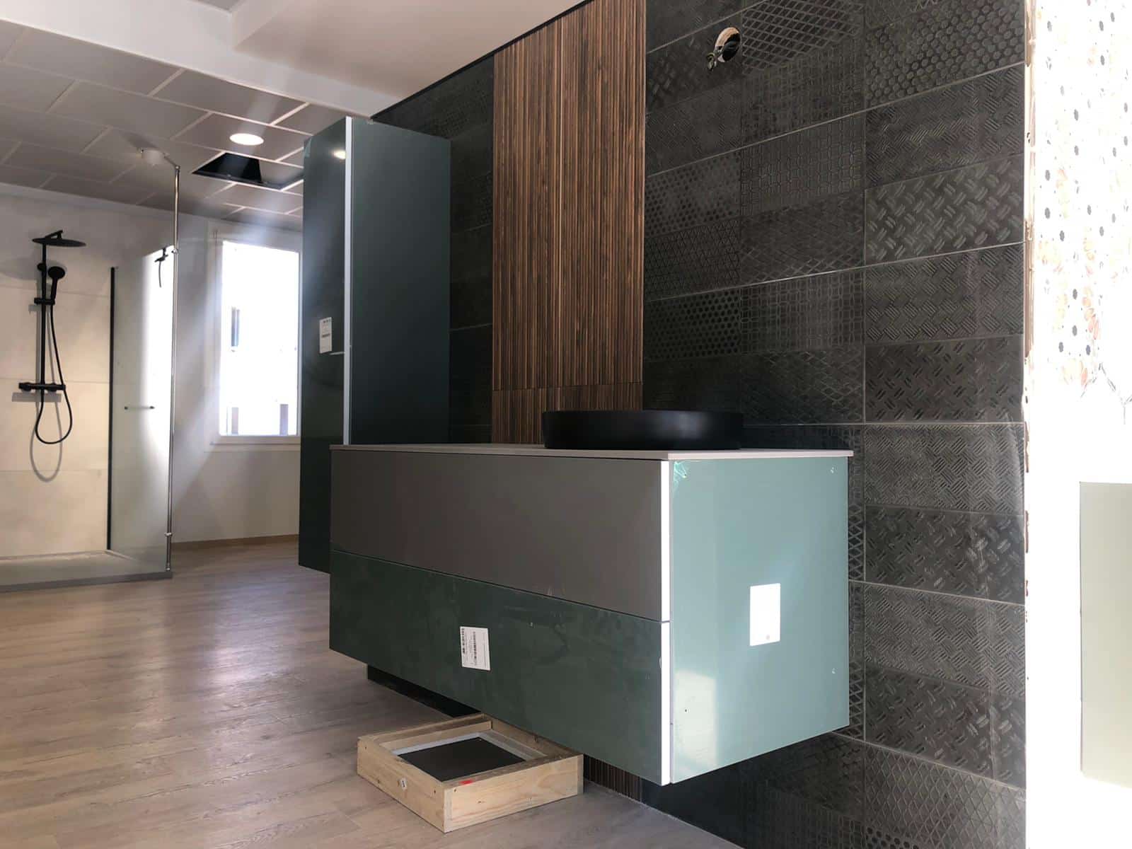 Intérieur d’un local technique : comptoir sombre avec face claire, mur en panneaux de métal texturé et colonne verticale en bois, sol en dalles, douchette et pomme de douche à gauche, caisson en bois au sol au premier plan.
