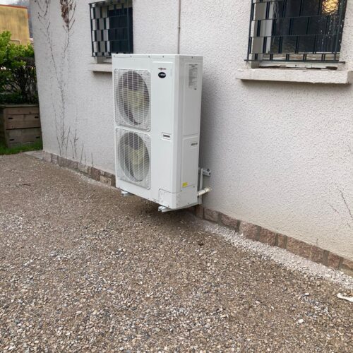 Un climatiseur extérieur blanc installé au pied d’un mur blanc, avec deux grilles de ventilation et deux fenêtres protégées par des grilles métalliques.