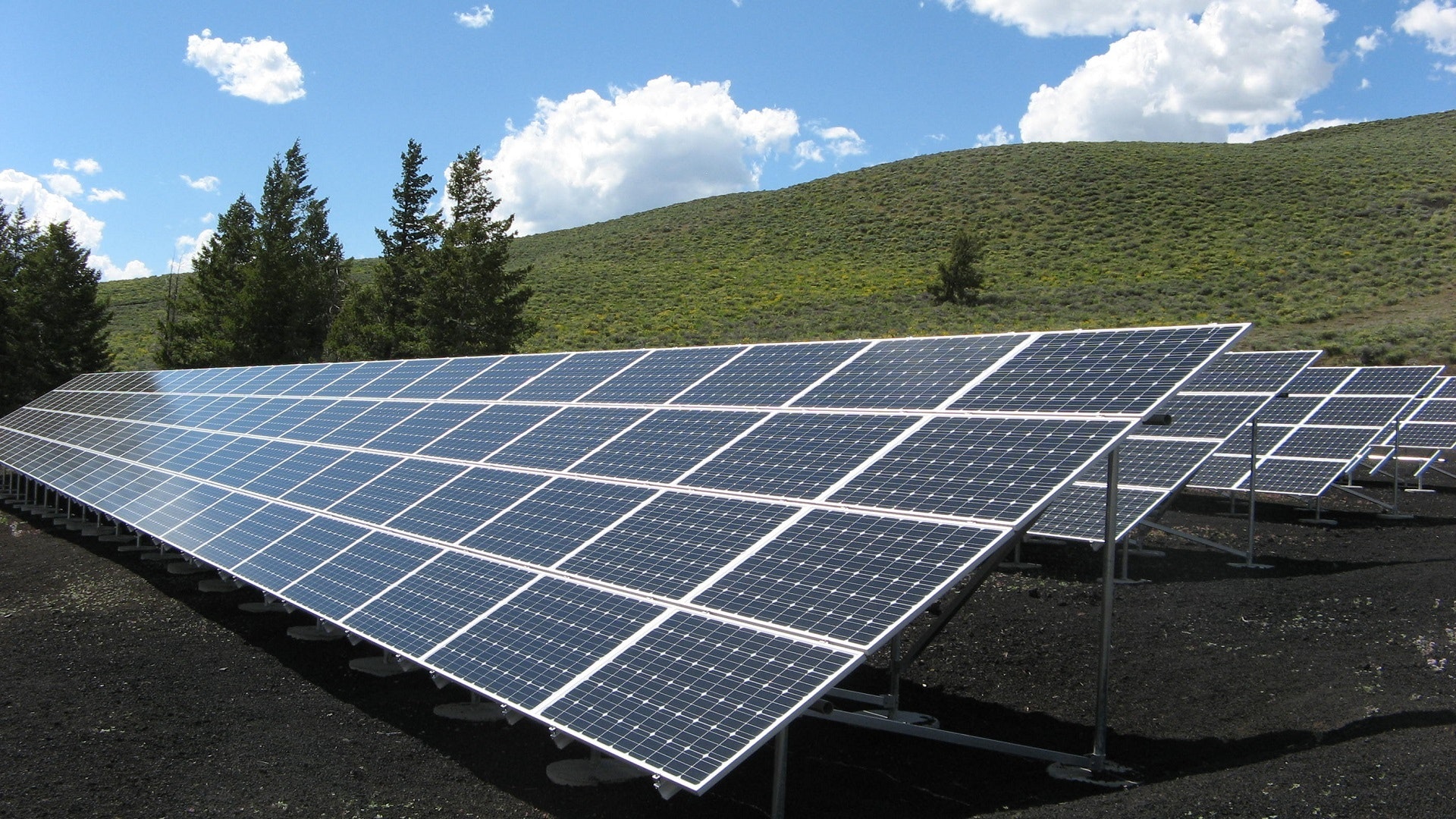 Batterie de panneaux solaires photovoltaïques installés au sol sur une pente, avec collines verdoyantes et ciel bleu nuageux en arrière-plan.