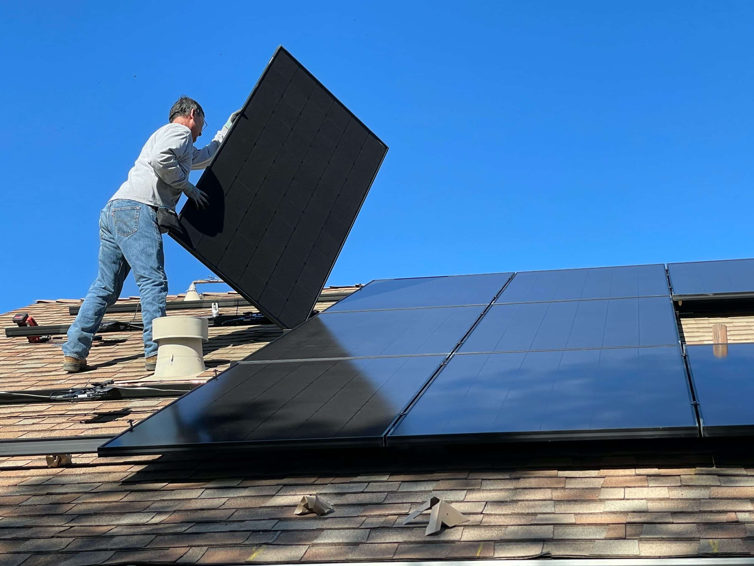 Un homme installe un panneau solaire noir sur un toit, par un ciel bleu, en soulevant le panneau avec ses mains.