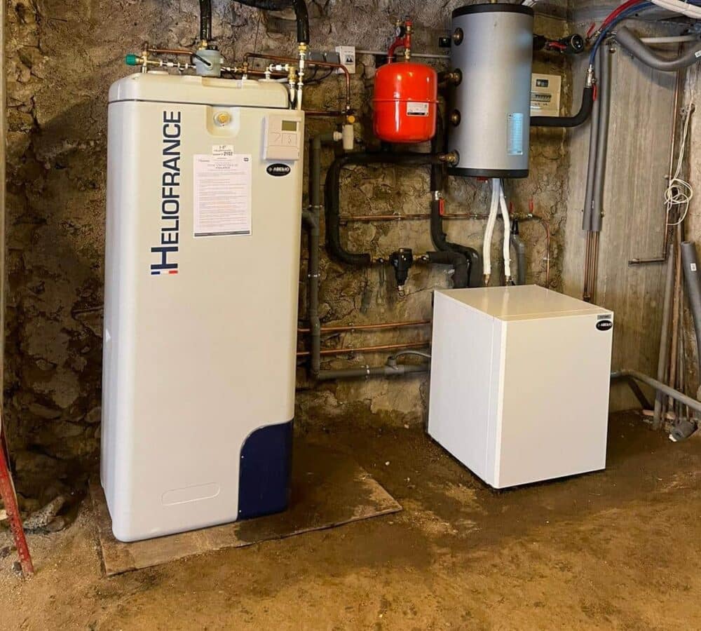 Local technique avec une chaudière murale blanc/bleu « HILOFRANCE » reliée à un réseau de tuyauteries, un réservoir cylindrique vertical, un ballon rouge et un coffret blanc sur fond de mur en pierre.