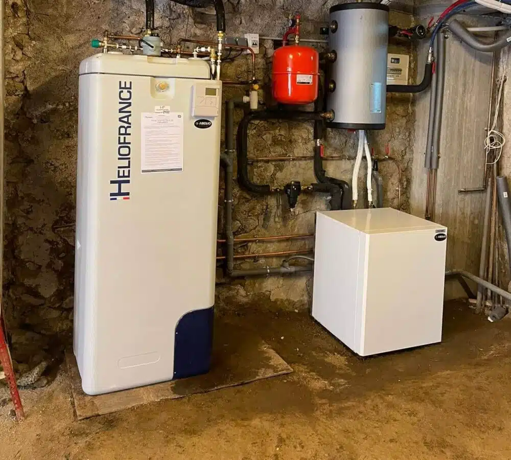 Local technique avec une chaudière murale blanc/bleu « HILOFRANCE » reliée à un réseau de tuyauteries, un réservoir cylindrique vertical, un ballon rouge et un coffret blanc sur fond de mur en pierre.