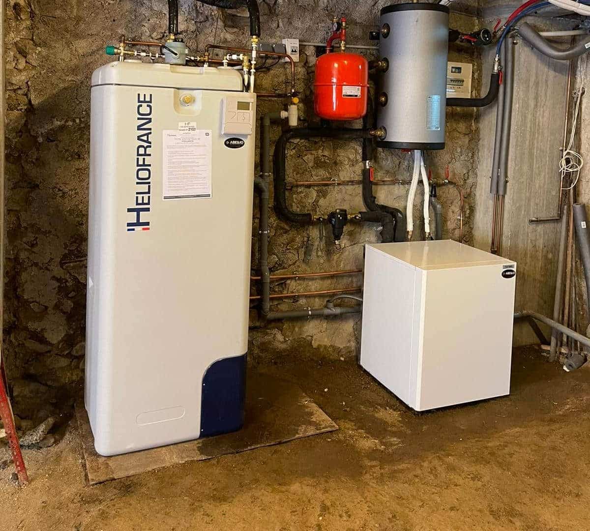 Local technique avec une chaudière murale blanc/bleu « HILOFRANCE » reliée à un réseau de tuyauteries, un réservoir cylindrique vertical, un ballon rouge et un coffret blanc sur fond de mur en pierre.