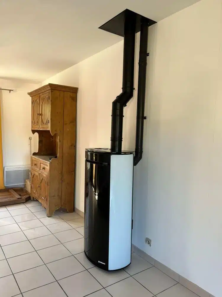 Poêle à granulés noir et blanc avec conduit de fumée installé dans une pièce, à côté d’un meuble en bois et du carrelage au sol.