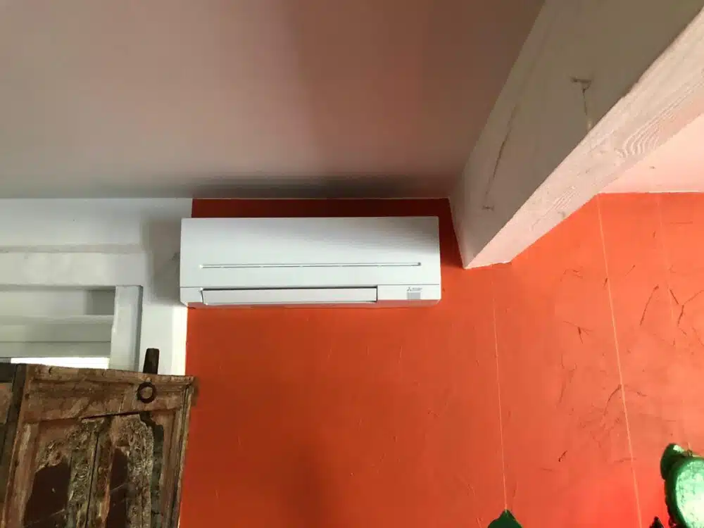 Climatisation murale blanche installée au-dessus d’un panneau orange, avec tuyaux visibles au plafond.