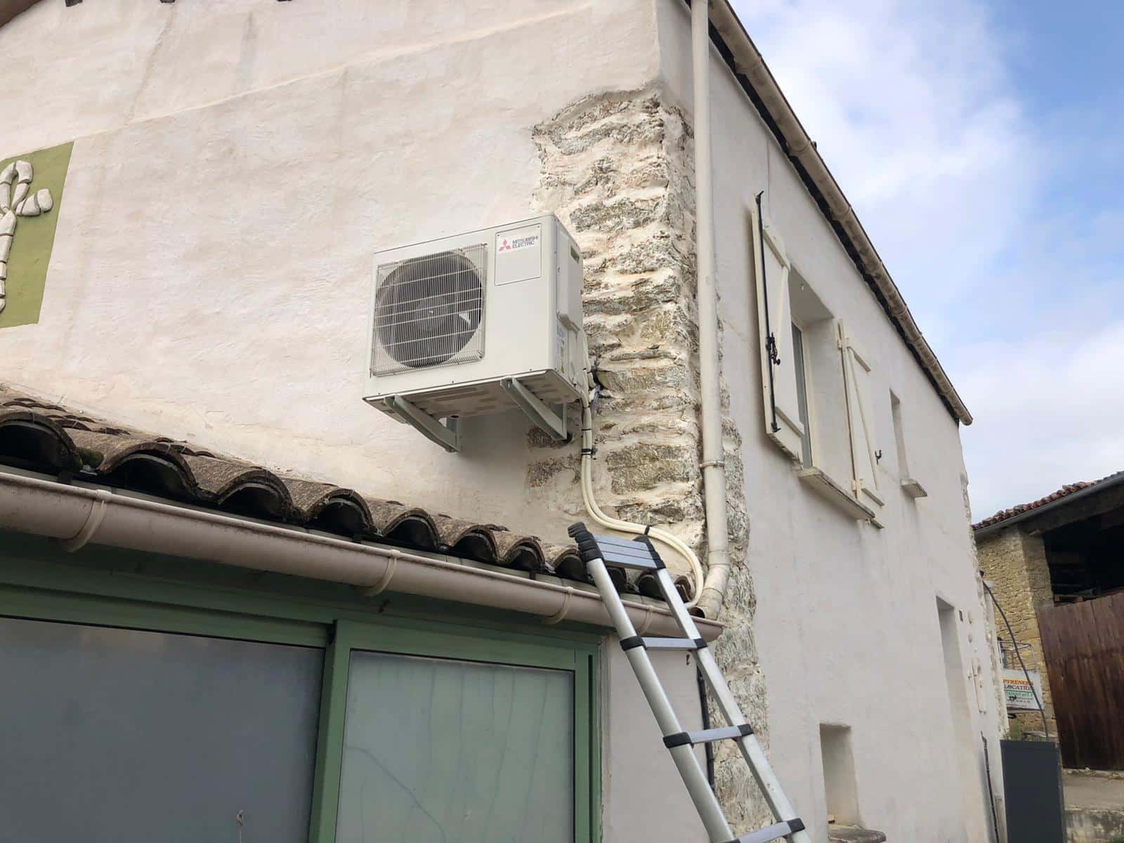 Climatiseur extérieur installé sur le mur d’une maison en pierre, avec tuyauteries et échelle devant, sous un ciel bleu.