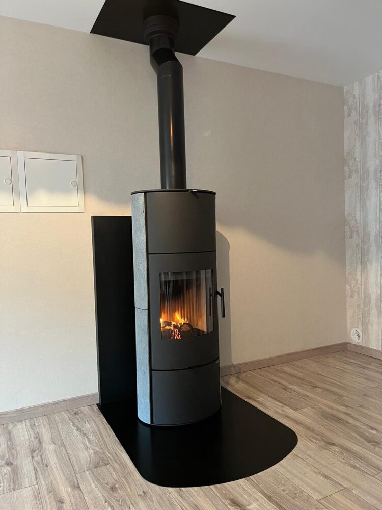 Poêle à bois noir allumé, avec cheminée sortant du plafond, installé dans un coin intérieur avec sol en parquet gris et mur clair.