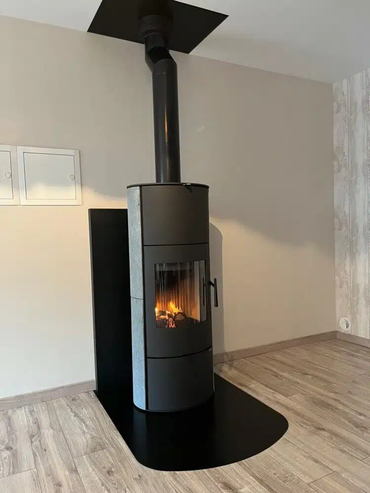 Poêle à bois noir allumé, avec cheminée sortant du plafond, installé dans un coin intérieur avec sol en parquet gris et mur clair.