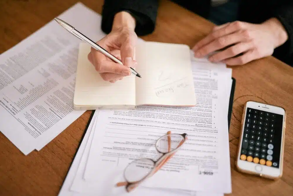 Mains écrivant sur des documents et livrets ouverts sur une table, avec une calculatrice et un smartphone à droite