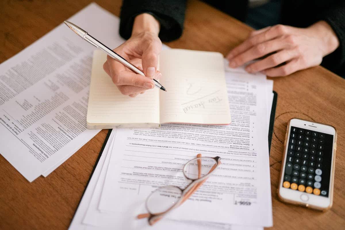 Mains écrivant sur des documents et livrets ouverts sur une table, avec une calculatrice et un smartphone à droite