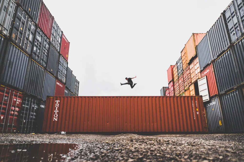 Un homme en saut acrobatique entre deux rangées de conteneurs maritimes, sur fond de ciel gris, devant un mur de conteneurs rouge.