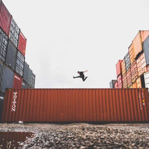 Un homme en saut acrobatique entre deux rangées de conteneurs maritimes, sur fond de ciel gris, devant un mur de conteneurs rouge.