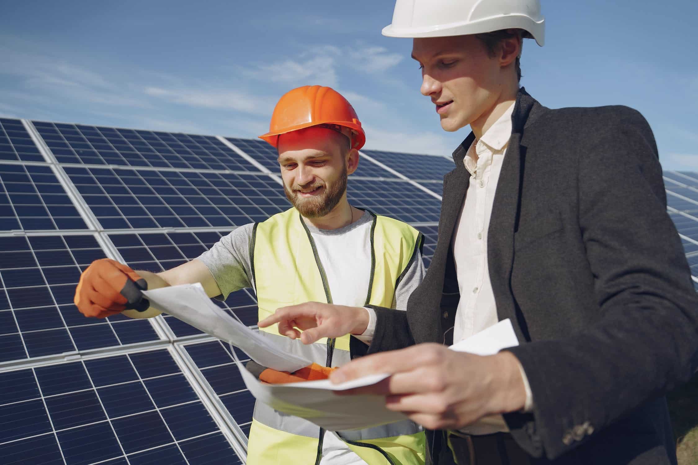 Deux techniciens en combinaison discutent et consultent des documents sur un toit équipé de panneaux solaires, sous un ciel dégagé.