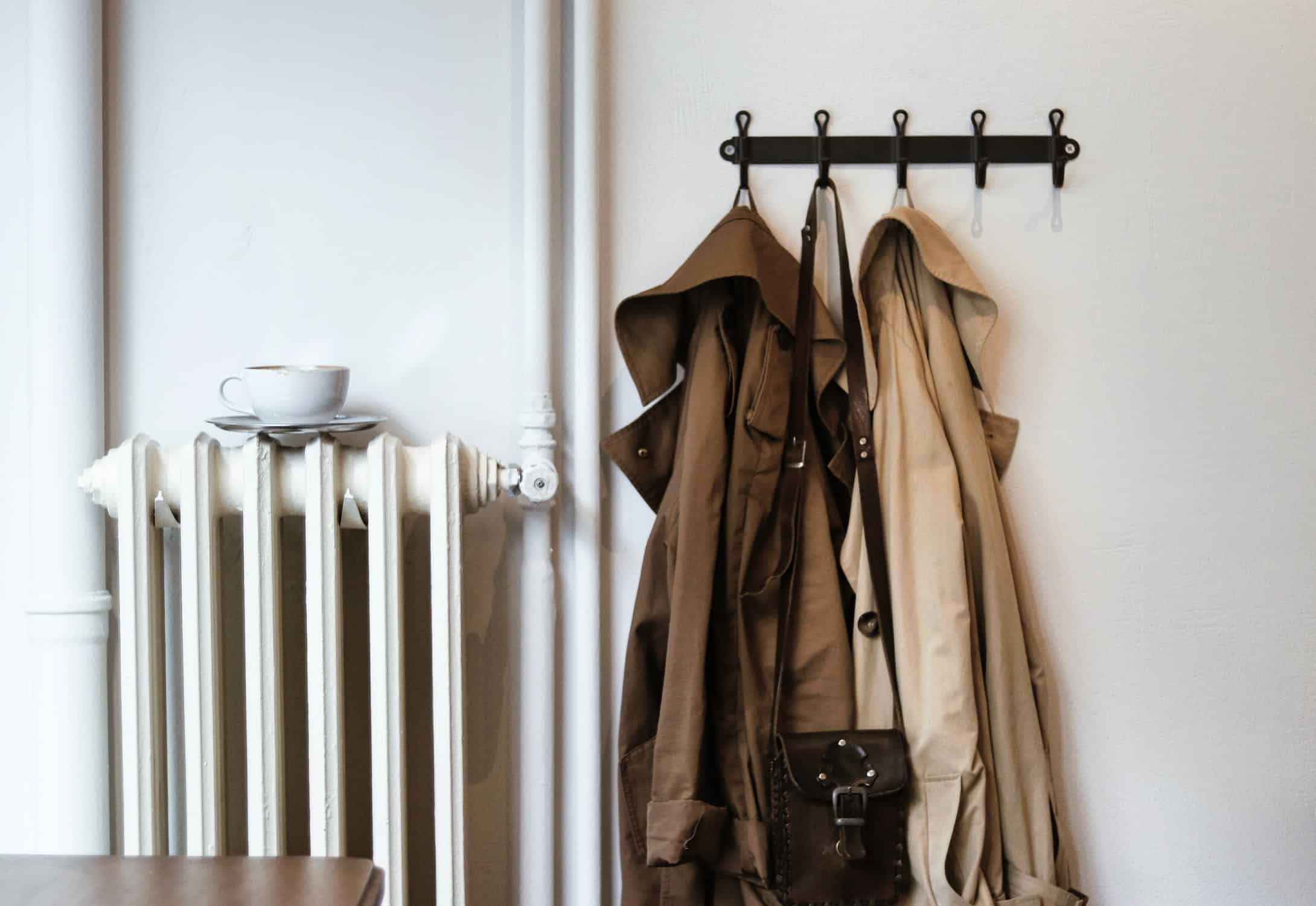 Manteaux beige et brun suspendus sur un porte-manteaux noir au mur, à côté d’un radiateur blanc et d’une tasse posée sur une étagère.