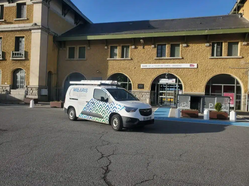 Véhicule de service blanc à motif géométrique stationné devant la gare de La Tour de Carol – Enveitg, entrée avec panneaux de bienvenue.