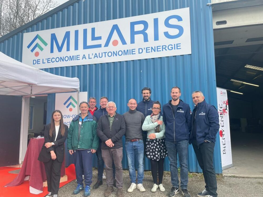 Photo de groupe devant un bâtiment bleu avec l’enseigne « MILARIS » et le slogan « De l’économie à l’autonomie d’énergie », plusieurs personnes posant devant une tente et un kakemono.