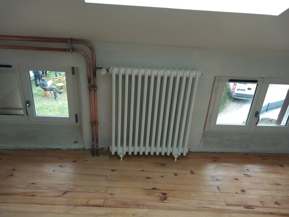 Pièce en cours d’aménagement avec sol en planches, radiateur blanc central, tuyaux cuivre au mur et fenêtres laissant voir l’extérieur, dont une personne assise dehors.