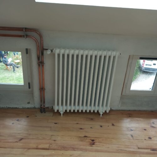 Pièce en cours d’aménagement avec sol en planches, radiateur blanc central, tuyaux cuivre au mur et fenêtres laissant voir l’extérieur, dont une personne assise dehors.