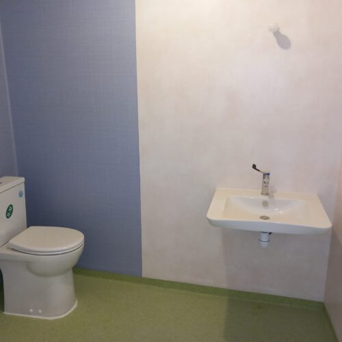 Salle de bain avec WC blanc à gauche et lavabo blanc à droite, robinet chromé et gel hydroalcoolique suspendu au mur.