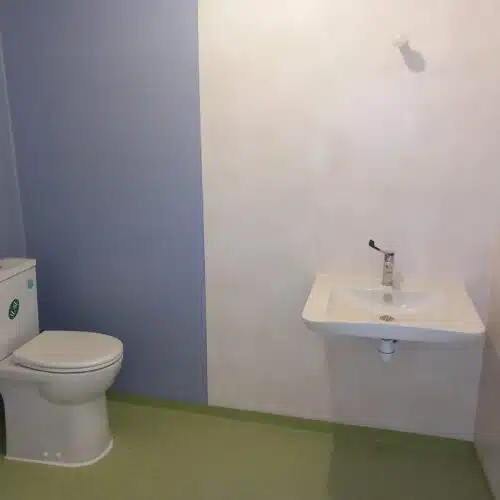 Salle de bain avec WC blanc à gauche et lavabo blanc à droite, robinet chromé et gel hydroalcoolique suspendu au mur.