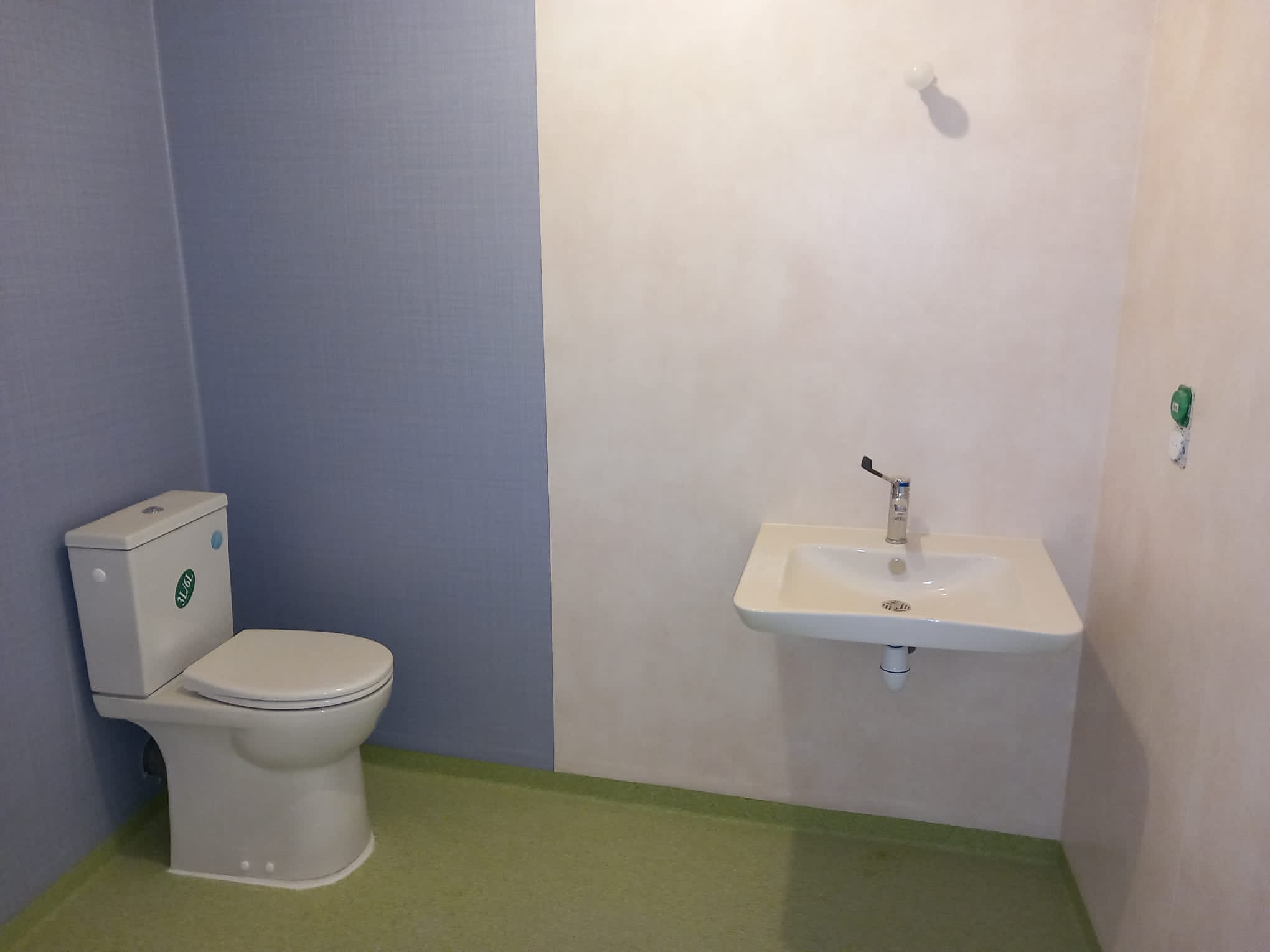 Salle de bain avec WC blanc à gauche et lavabo blanc à droite, robinet chromé et gel hydroalcoolique suspendu au mur.