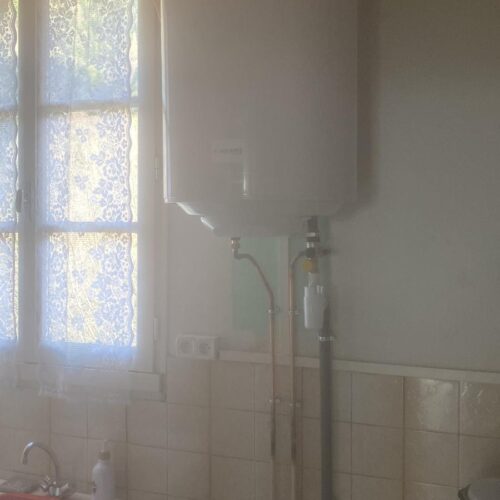 Pièce blanche avec tuyauterie apparente et chauffe-eau mural, fenêtre avec rideau à motifs au fond et lavabo sur le côté gauche, avec carrelage mural clair.
