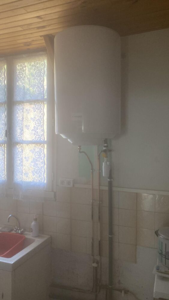 Pièce blanche avec tuyauterie apparente et chauffe-eau mural, fenêtre avec rideau à motifs au fond et lavabo sur le côté gauche, avec carrelage mural clair.