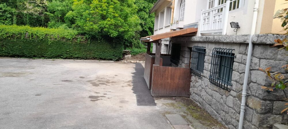 Cour pavée devant une maison, avec mur en pierres, grilles métalliques et volets, bordée par une végétation dense et des arbres verts.