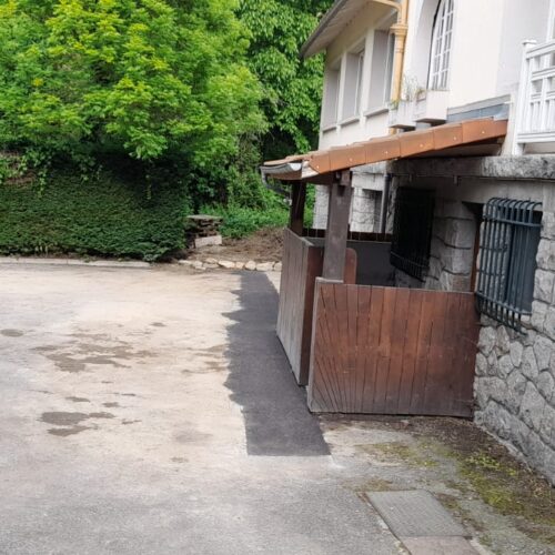 Cour pavée devant une maison, avec mur en pierres, grilles métalliques et volets, bordée par une végétation dense et des arbres verts.