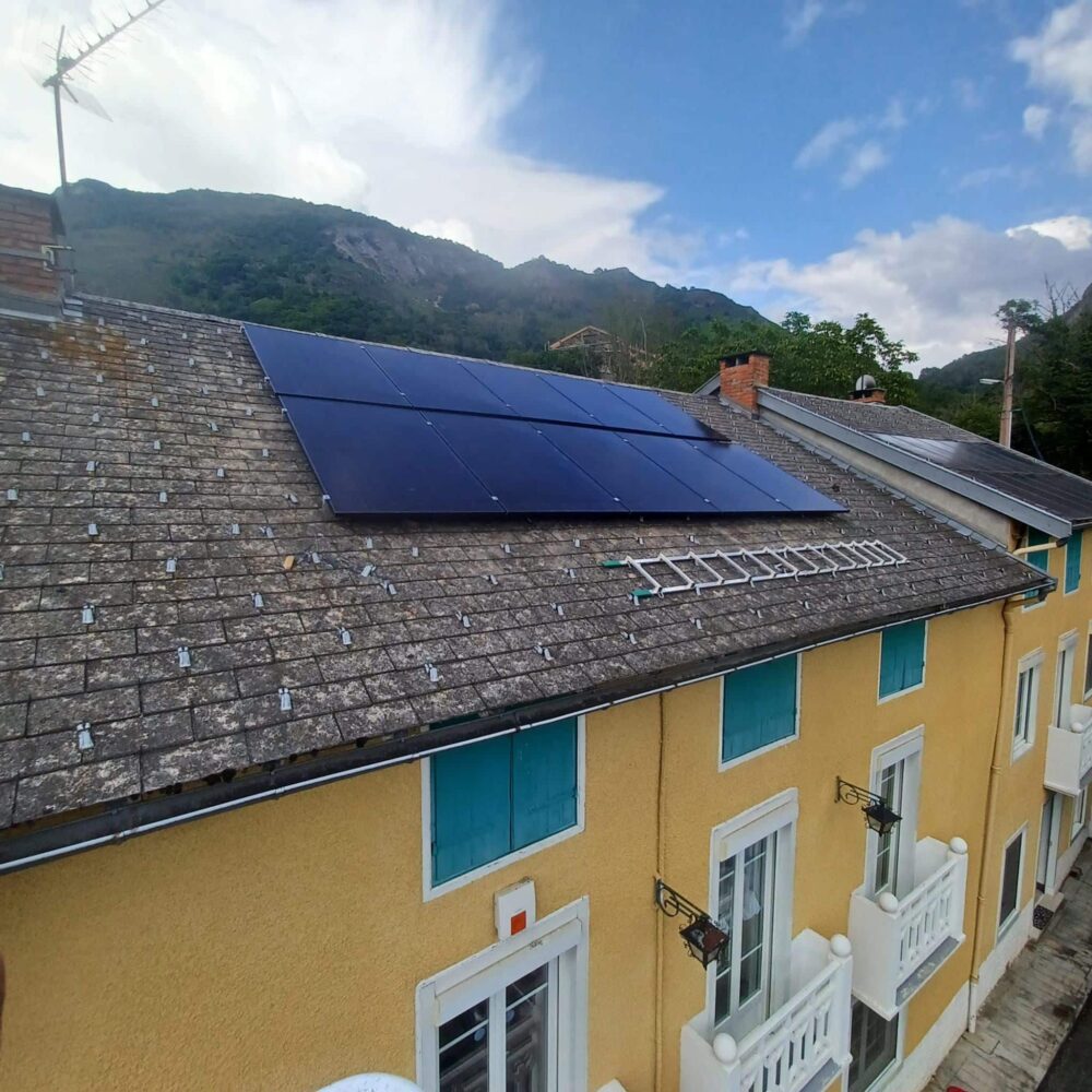 Toit d’une maison recouvert de panneaux solaires bleus, sur fond de collines et de ciel partiellement nuageux.