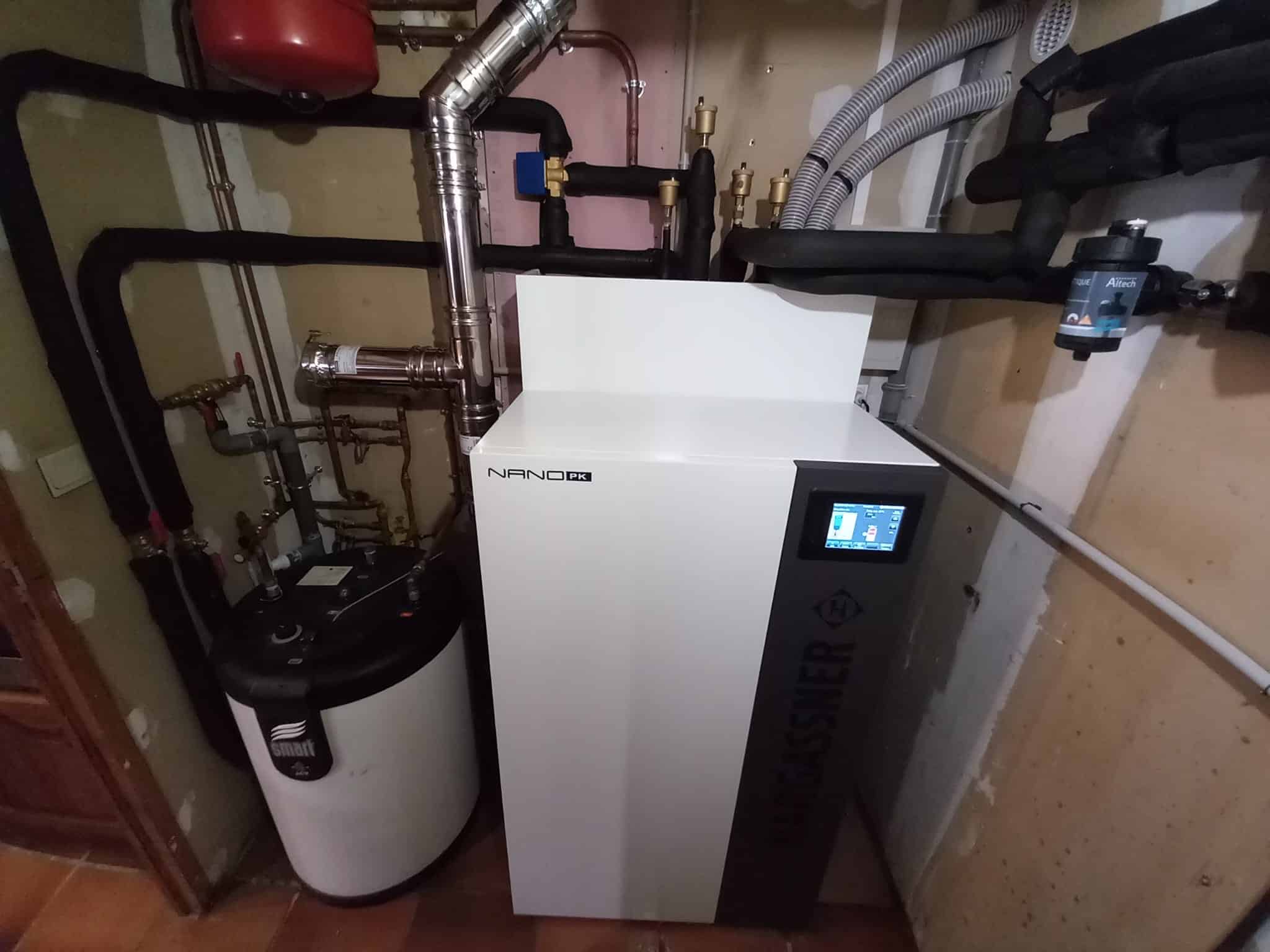 Local technique avec une chaudière industrielle blanche et son écran de contrôle, entourée de tuyauteries métalliques, vannes et conduits de ventilation, avec un ballon rouge au plafond.