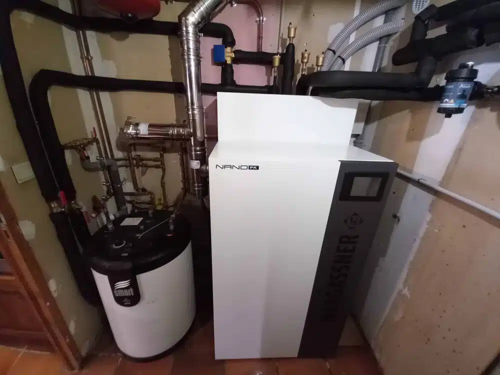 Local technique avec chaudière NANO PKI murale, tuyauteries métalliques et conduits isolés, et régulateur sur pied avec écran.