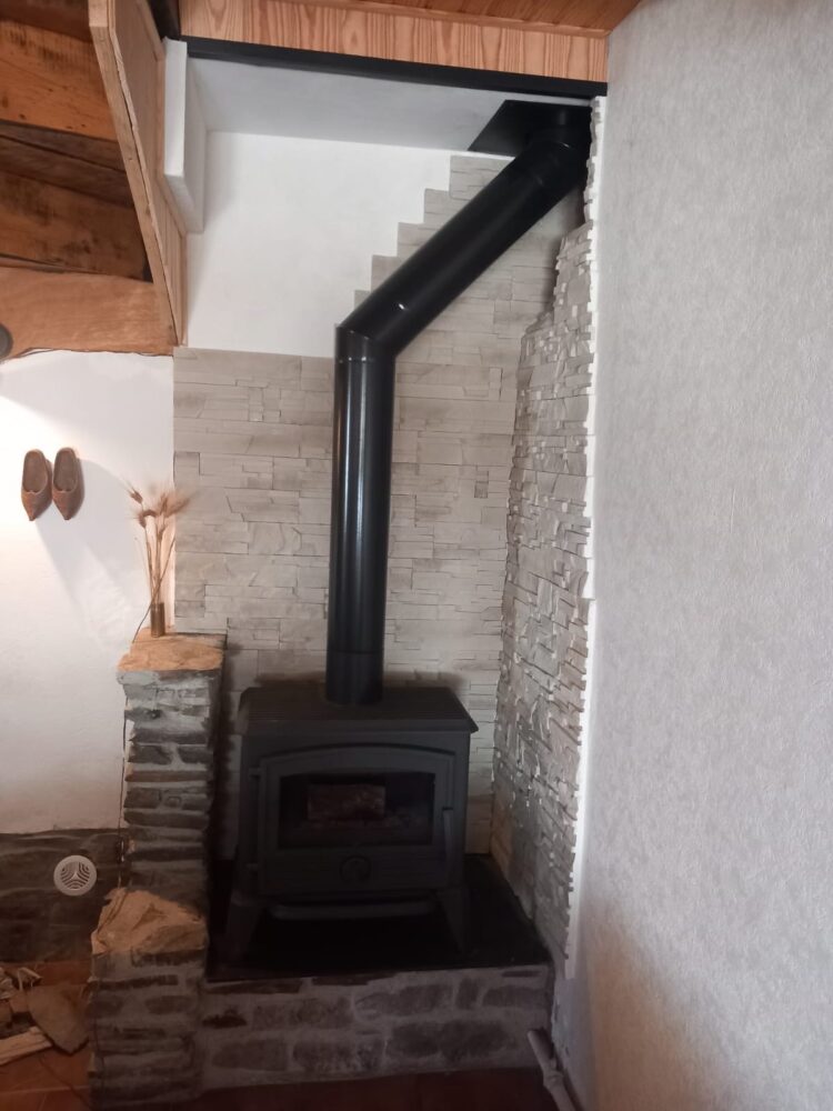 Poêle à bois noir encastré dans un mur de briques, avec conduit vertical et conduit courbé menant au plafond.