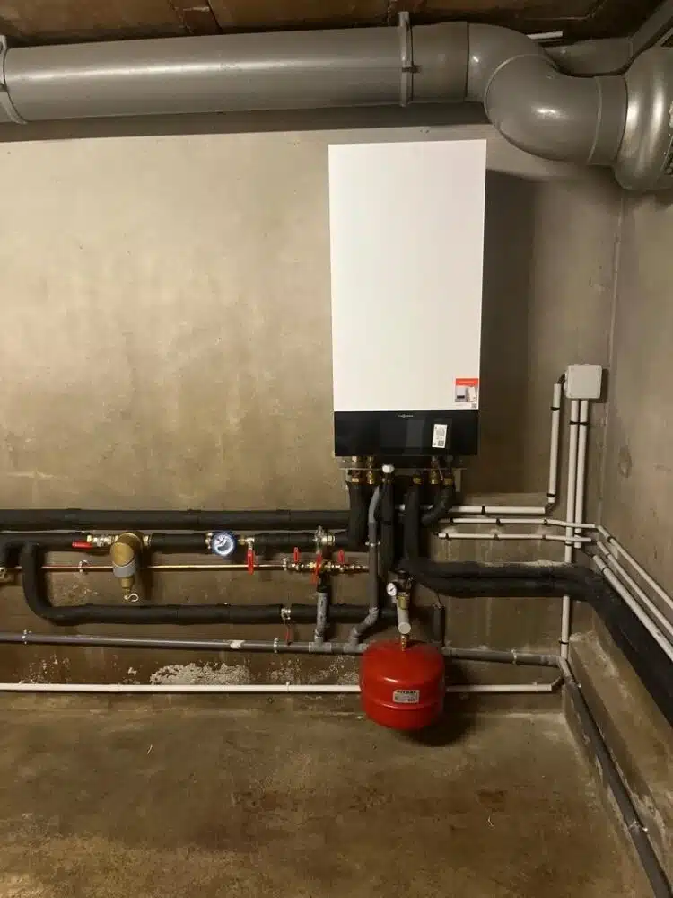 Local technique avec chaudière murale blanche et tuyauteries industrielles en acier, vannes rouges et bouteille rouge au sol, dans une gaine béton avec conduits de ventilation.