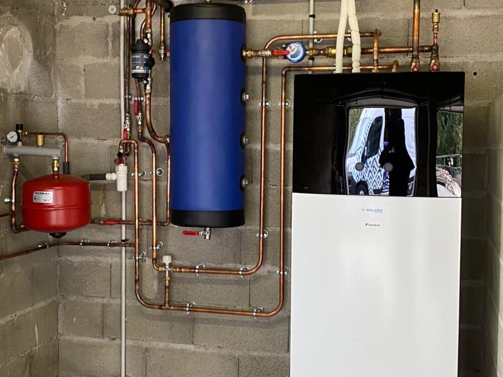 Local technique en sous-sol avec tuyauteries cuivre, réservoir bleu cylindrique, filtre rouge et unité de chauffage/climatisation blanche, posée contre un mur en béton.