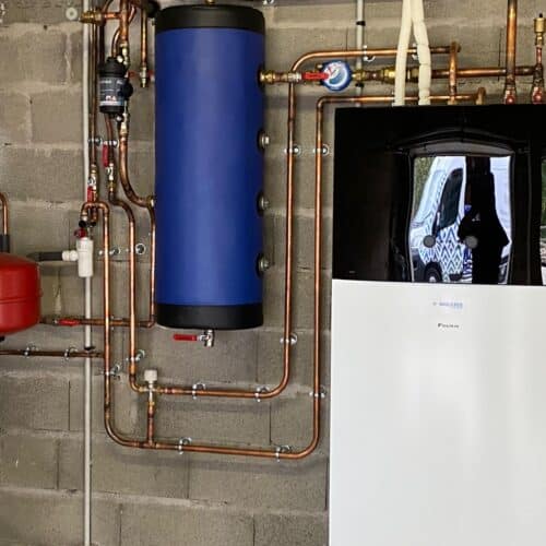 Local technique en sous-sol avec tuyauteries cuivre, réservoir bleu cylindrique, filtre rouge et unité de chauffage/climatisation blanche, posée contre un mur en béton.