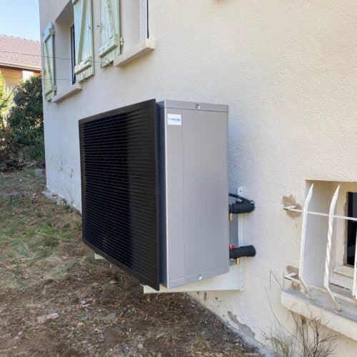 Un groupe extérieur de climatisation sur le mur d’une maison, avec conduits raccordés et grilles de ventilation, au pied d’une façade blanche.