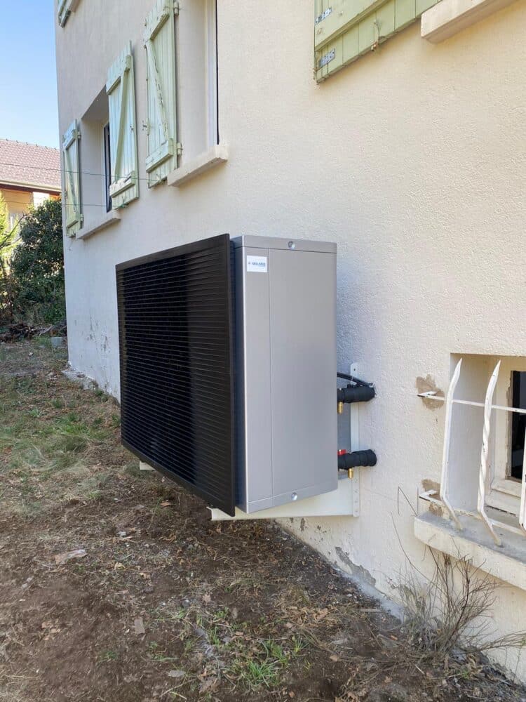 Un groupe extérieur de climatisation sur le mur d’une maison, avec conduits raccordés et grilles de ventilation, au pied d’une façade blanche.
