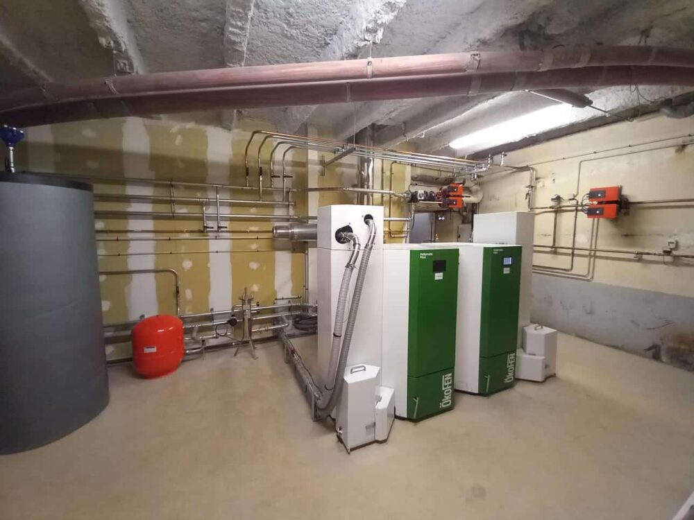 Local technique d’une chaufferie : tuyauteries en acier, conduits au plafond et deux chaudières vertes avec des unités blanches, ainsi qu’un ballon rouge et des échangeurs, dans une pièce bétonnée.