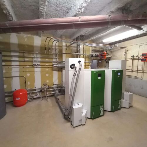 Local technique d’une chaufferie : tuyauteries en acier, conduits au plafond et deux chaudières vertes avec des unités blanches, ainsi qu’un ballon rouge et des échangeurs, dans une pièce bétonnée.