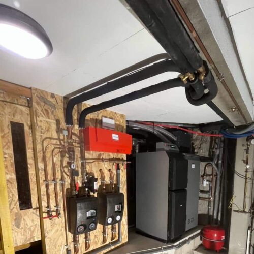 Local technique d’une chaufferie : tuyauteries, vannes et chaudière noire sur un mur en panneaux, avec conduites isolées, câblage et petit réservoir rouge au sol.