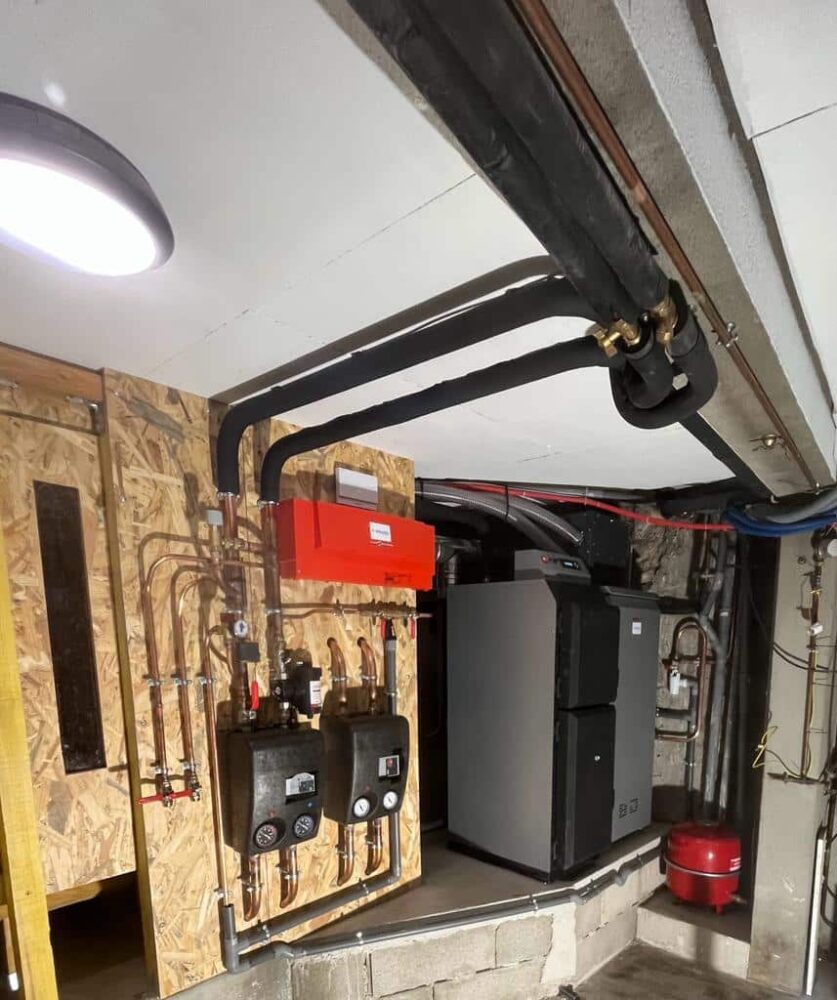 Local technique d’une chaufferie : tuyauteries, vannes et chaudière noire sur un mur en panneaux, avec conduites isolées, câblage et petit réservoir rouge au sol.