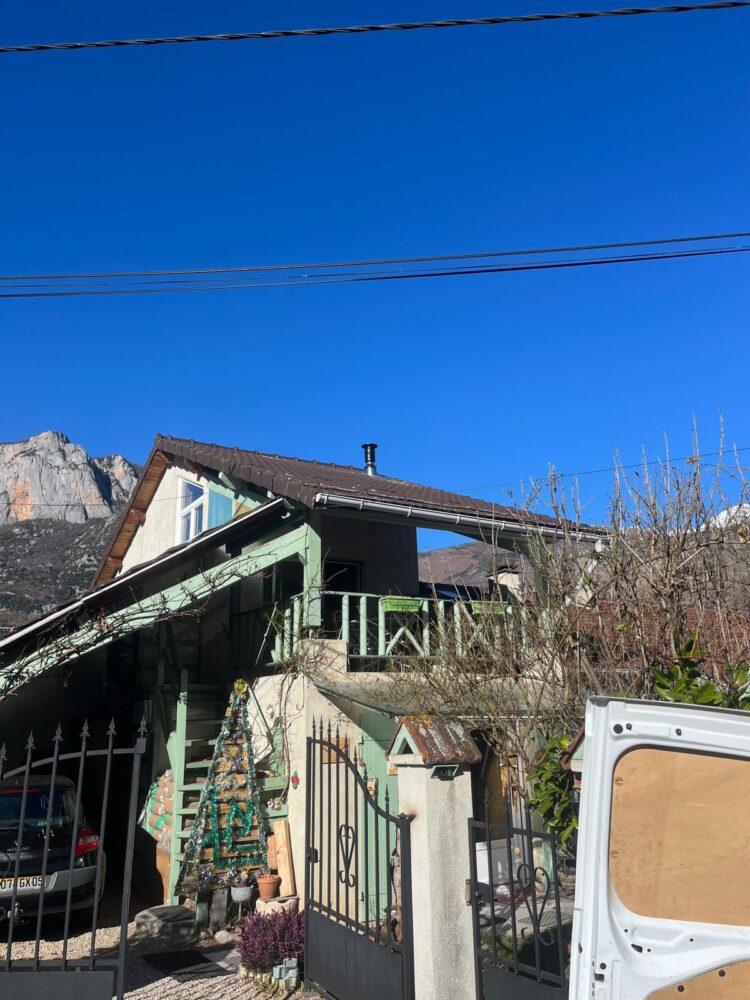 Façade d’une maison verte avec toit en tuiles, contrevent et cheminée, sous un ciel bleu, avec jardin devant et un petit sapin décoré près d’une clôture en fer.