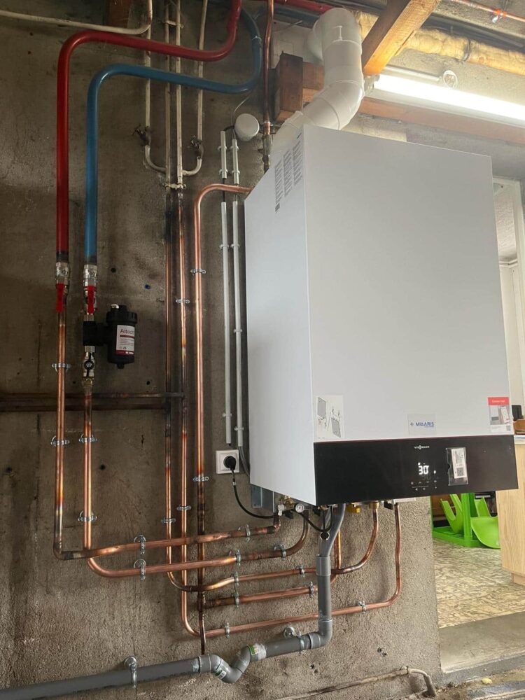 Chaudière murale blanche avec écran de commande, raccordée à un réseau de tuyauteries en cuivre et conduits, dans une chaufferie avec murs en béton et câbles.