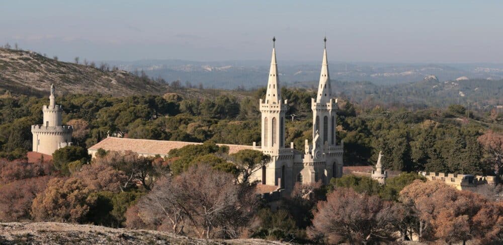 Vue aérienne d’une basilique blanche à deux tours pointues sur un relief boisé, avec un château à tourelle à gauche et des montagnes au loin.