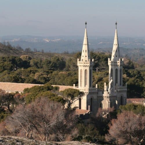 Vue aérienne d’une basilique blanche à deux tours pointues sur un relief boisé, avec un château à tourelle à gauche et des montagnes au loin.