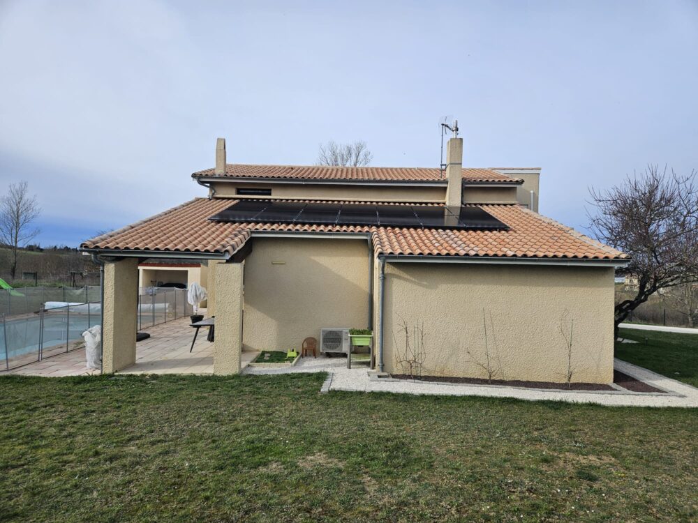 Façade d’une maison avec toit en tuiles, cheminée centrale et panneaux solaires sur la toiture, vue depuis le jardin, ciel bleu clair.